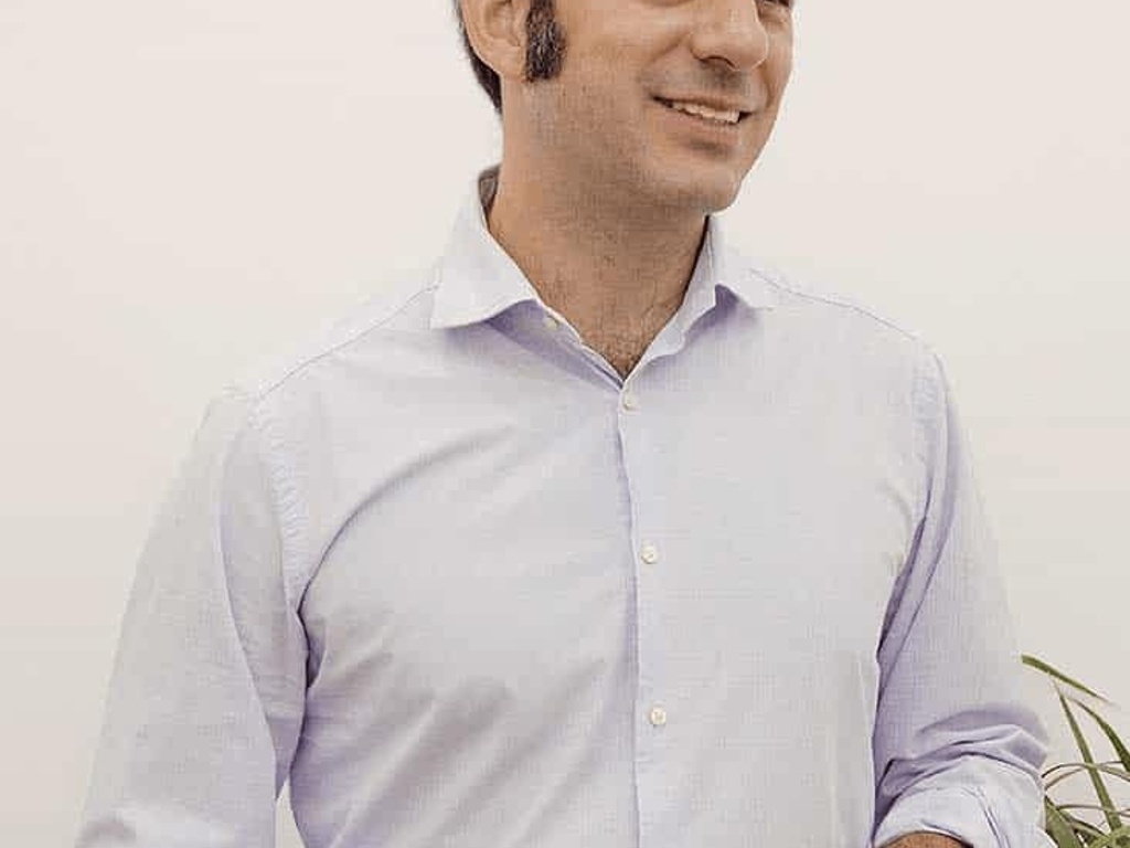 A man in a light blue shirt holding a white mug, smiling and looking to the side.