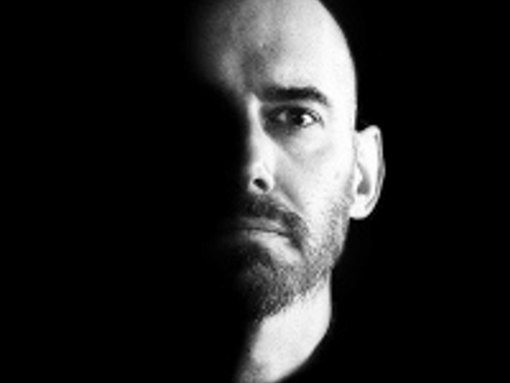 Black and white portrait of a bald man with a beard looking intently towards the camera, illuminated from one side.