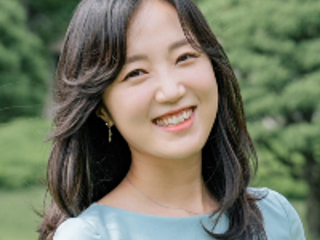 A smiling young woman with long hair in a light blue dress standing in a park-like setting.