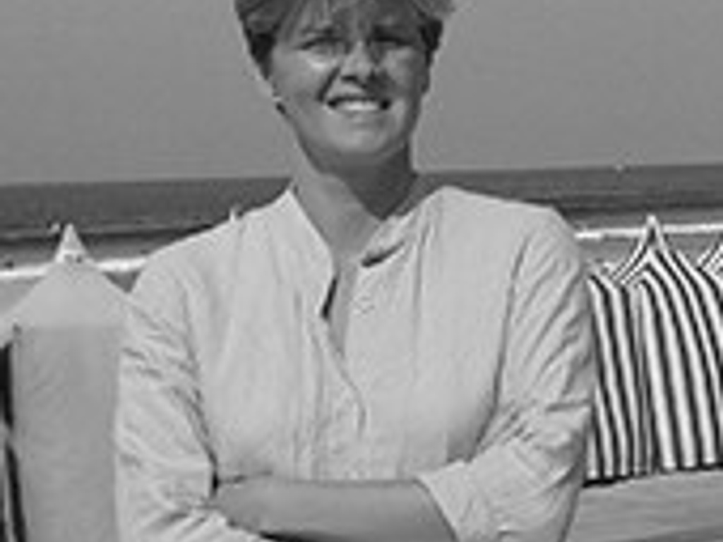 A black and white photo of a smiling woman with short hair, dressed in a light blouse, standing on a beach with a striped beach chair in the background.