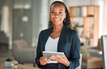 A smiling businesswoman holds a tablet while standing in a modern office.