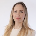 Portrait of a woman with long blonde hair wearing a light beige top, standing against a plain white background.