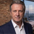 A serious-looking man in a suit poses in a modern office environment.
