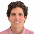 Portrait of a smiling young man with curly hair wearing a pink shirt, against a plain white background.