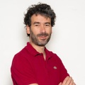 A man with curly hair and a beard is posing confidently with crossed arms.