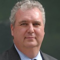 A professional older man with wavy, graying hair and a business suit posing in front of a green background.