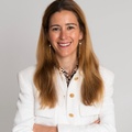 A smiling woman with long hair wearing a white blazer and earrings, standing with her arms crossed.