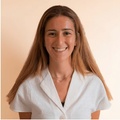 A portrait of a smiling woman with long hair wearing a white shirt against a light pink background.