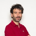 A man with curly hair and a beard is posing confidently with crossed arms.
