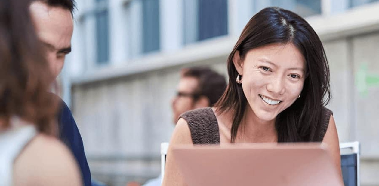 A woman smiling while looking at a laptop, engaging in a discussion with a man partially visible across the table.