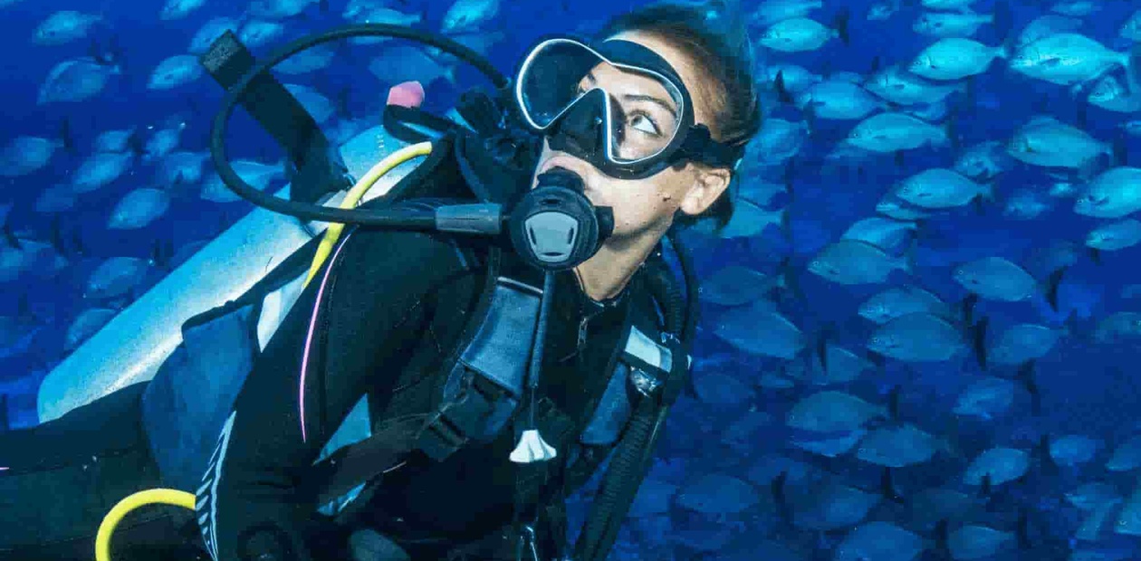 A female diver swims underwater surrounded by a school of small blue fish.