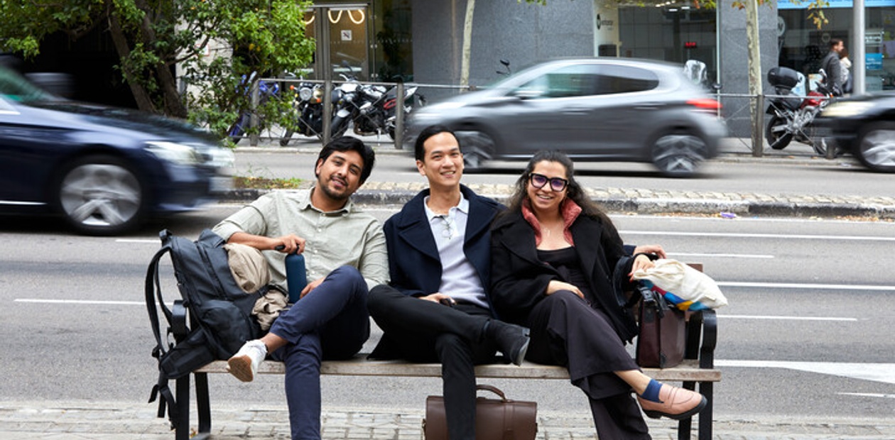 Three people sitting on a bench on a city sidewalk, smiling as traffic passes by.