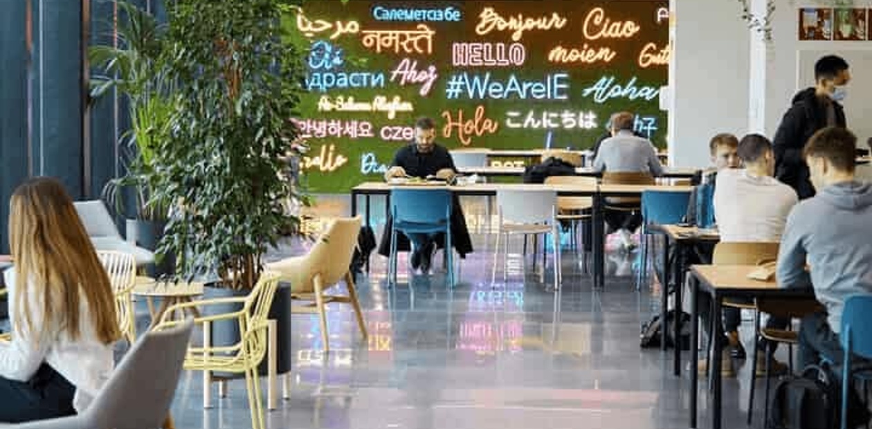 A modern and bustling cafeteria with people seated at tables, and a colorful wall with multi-language greetings.