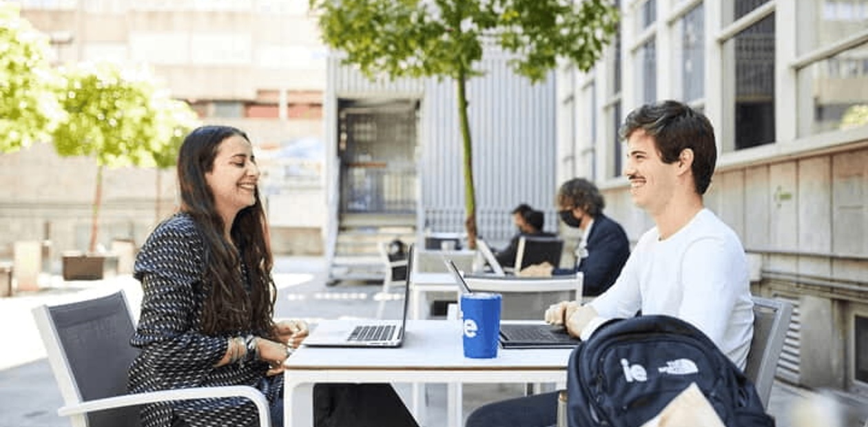 Two individuals, a man and a woman, are sitting at a table outdoors, having a conversation with laptops in front of them.