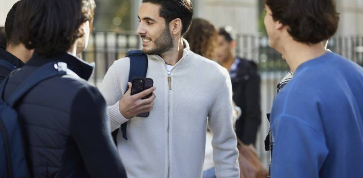 Three young men are having a conversation outdoors, with one showing something on his smartphone to the others.