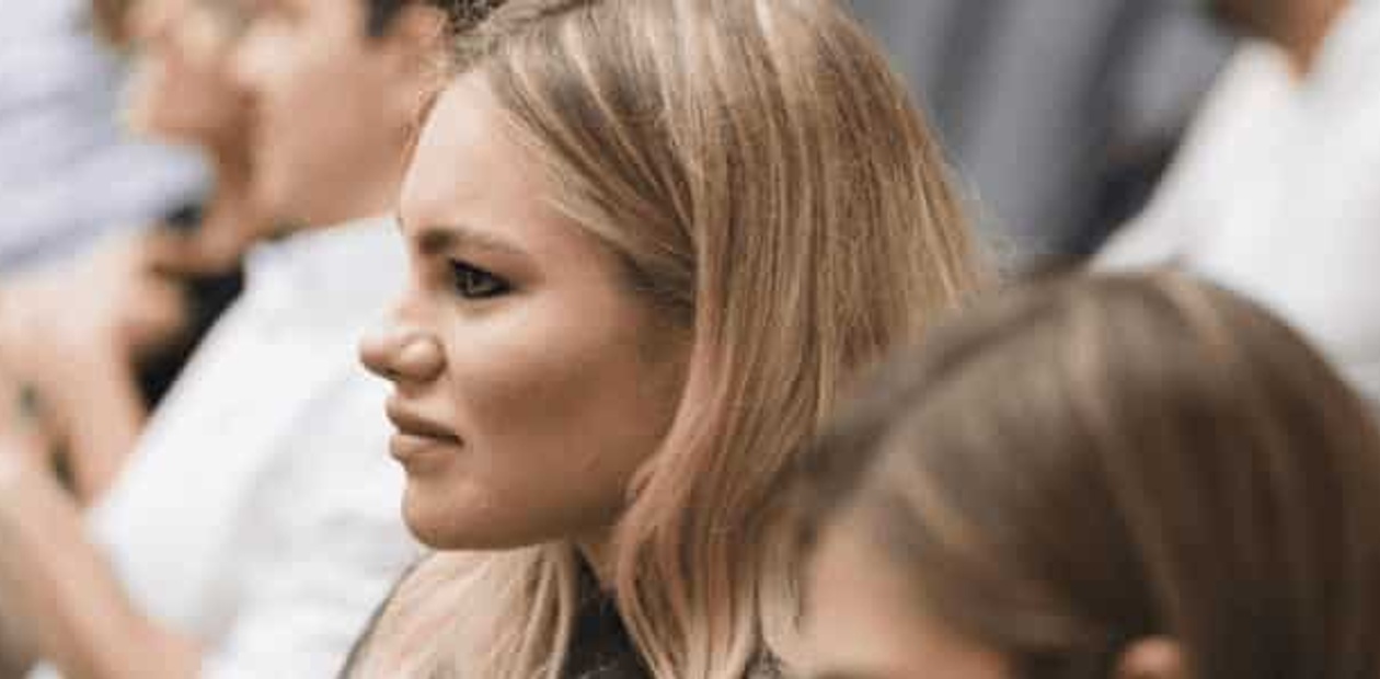 A young woman is sitting and listening attentively in a conference or seminar setting with other attendees in the background.