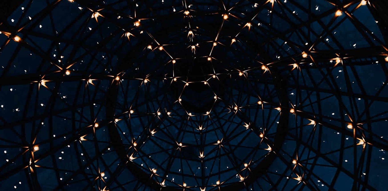 A view from inside a dome structure with a geometric framework illuminated by small lights against a twilight sky.