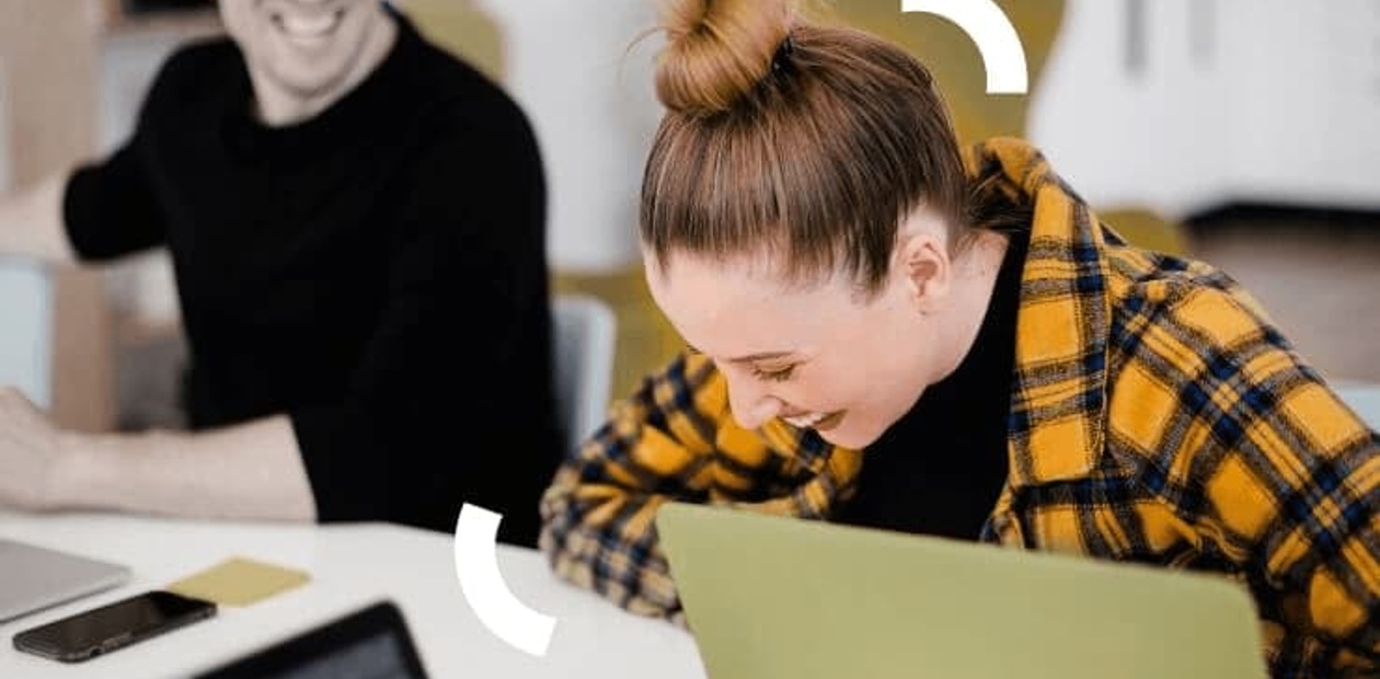 A woman and a man are smiling while working on laptops in a modern office environment.