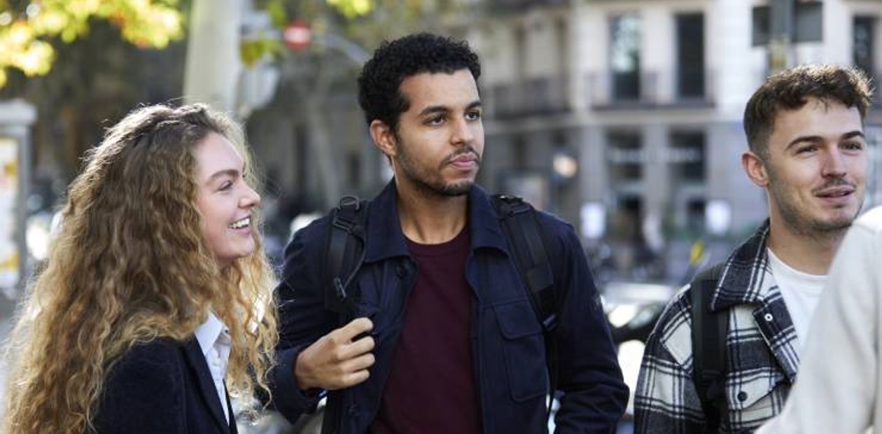 Three young adults are walking and smiling together on a city street lined with trees.