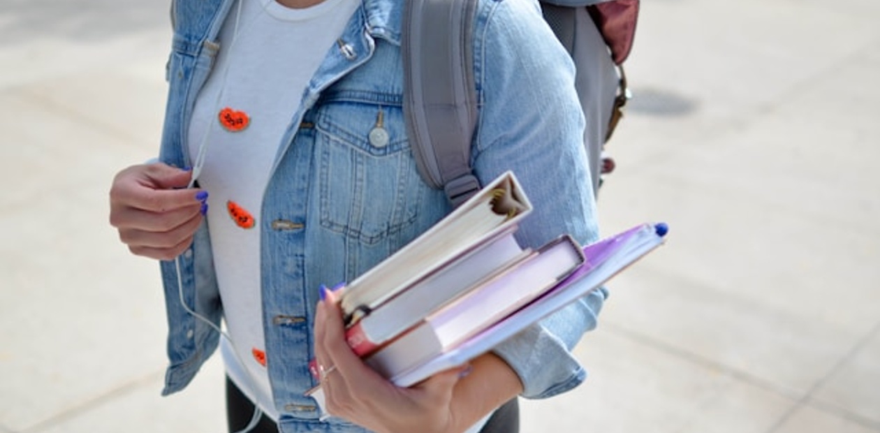 A person holding several books while wearing a denim jacket and backpack.