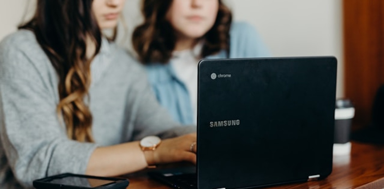 Two women are working on a laptop at a wooden table.