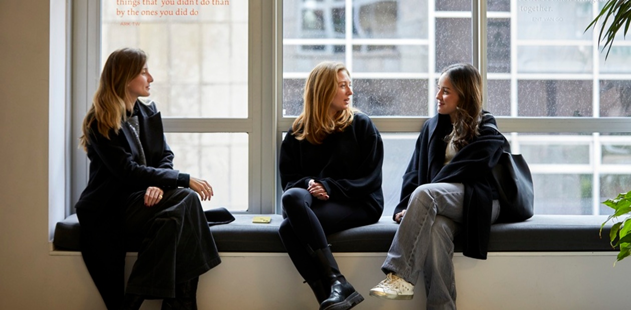 Three women sitting together in a modern space, discussing something while looking out a window.