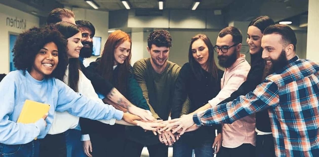 A group of diverse young adults are stacking hands together in a gesture of teamwork and unity in an office environment.