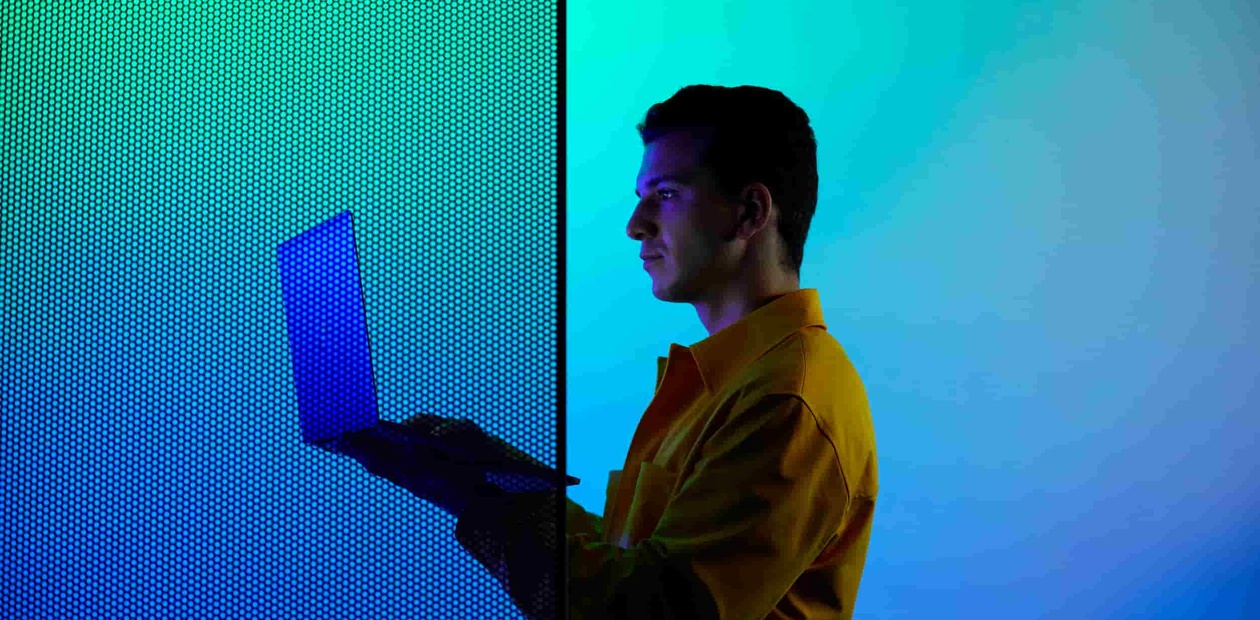 A man in a yellow jacket uses a laptop in front of a large blue glowing screen.