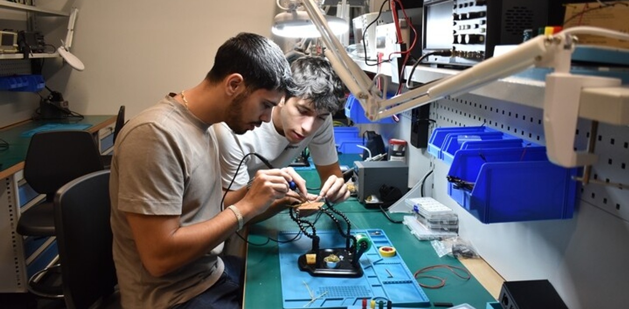 Two young men are collaboratively working on an electronic project at a workbench.