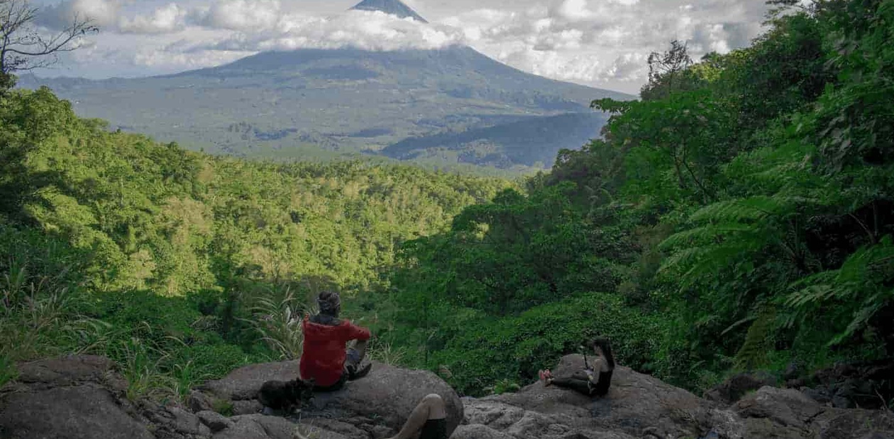 Two people sitting on rocks enjoying the view of a distant volcano surrounded by lush greenery.