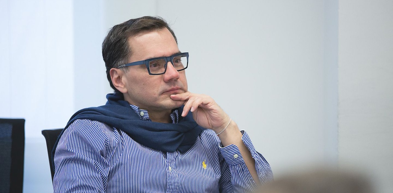 A man deep in thought wearing glasses and a sweater draped over his shoulders.