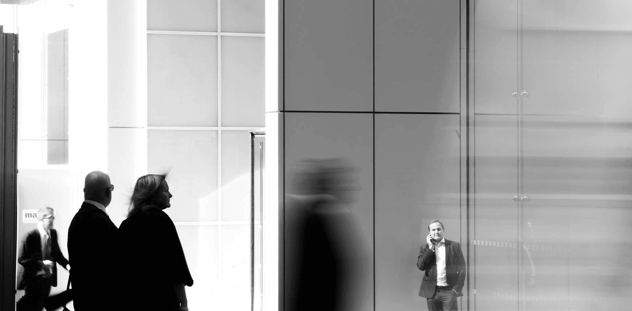 Black and white image of people standing and walking in a modern building with glass walls.