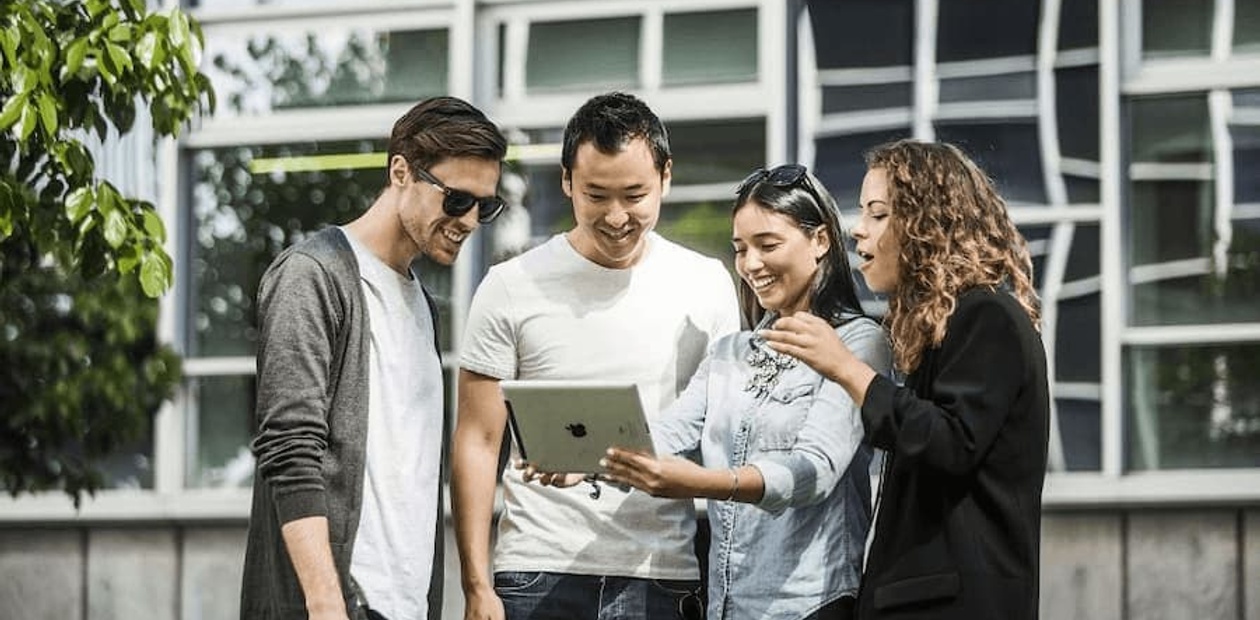 Four young adults, two men and two women, are standing outdoors and looking at a tablet together with expressions of engagement and happiness.