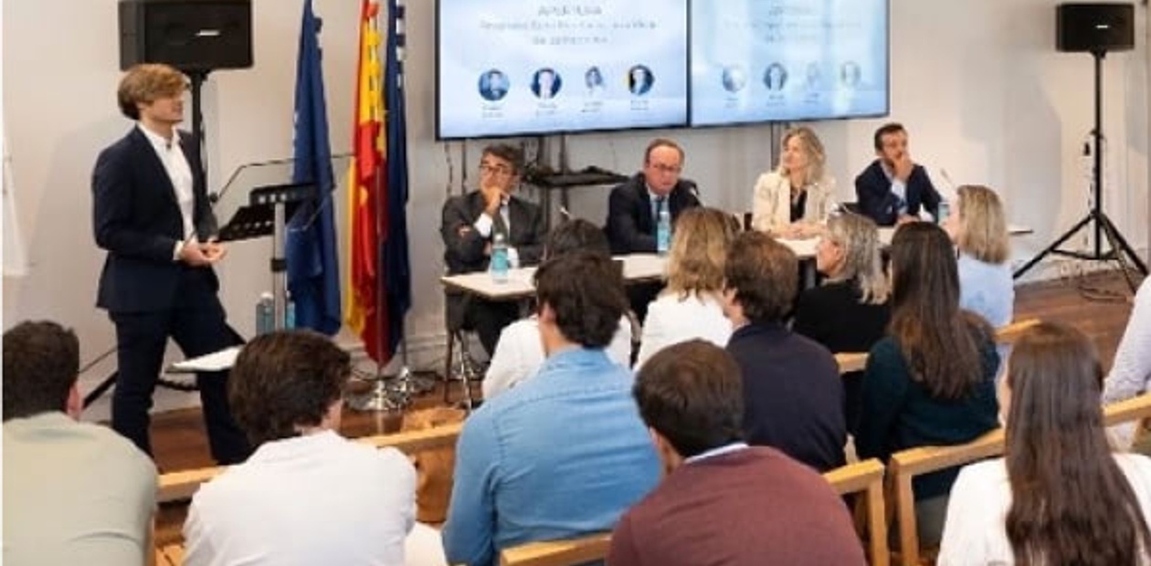 A young man speaks at a podium in a conference room with an audience and panelists watching him.