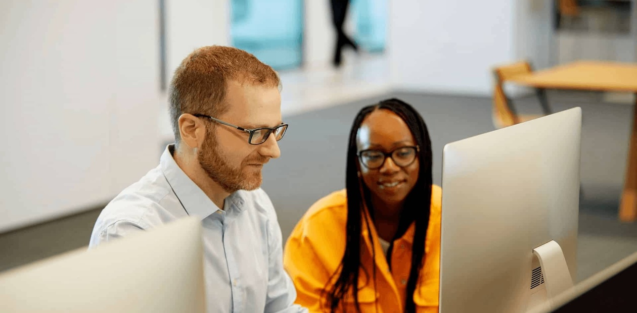 A man and a woman sitting at a desk, collaboratively working on computers in an office setting.
