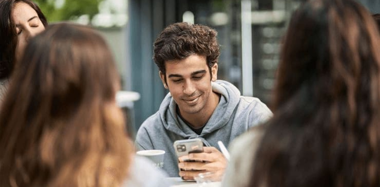 A young man smiling at his phone while sitting at a table with several friends around him outdoors.