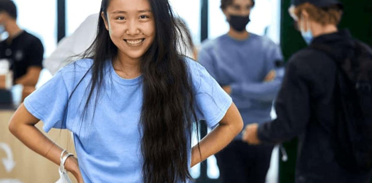 A smiling young woman in a blue t-shirt standing in a busy indoor environment.