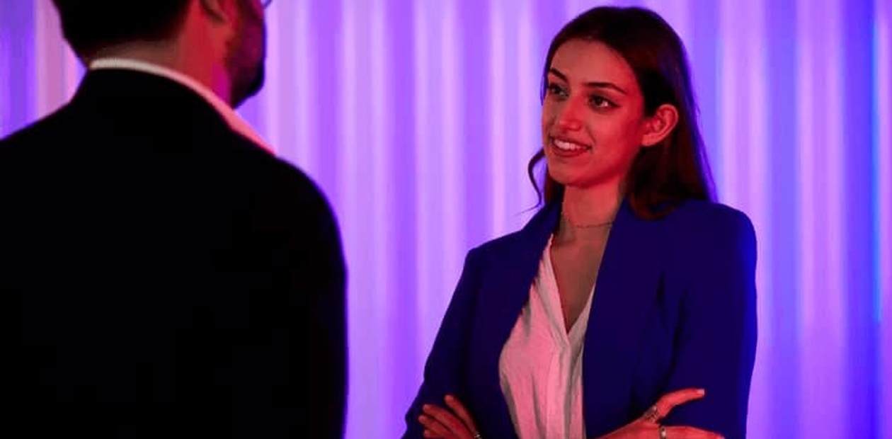 A woman in a blue blazer is talking to a man in a dark suit, illuminated by purple lighting in the background.