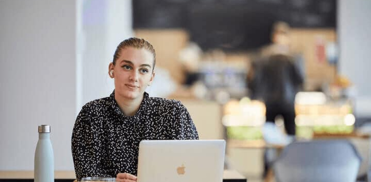 A woman sits at a table in a cafe with a laptop and a coffee cup in front of her.