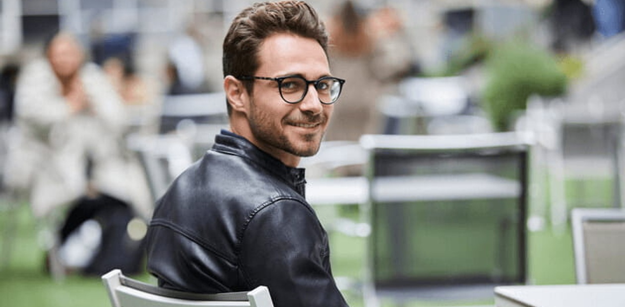 A man in a leather jacket and glasses smiling and sitting at a table in an outdoor cafe setting.