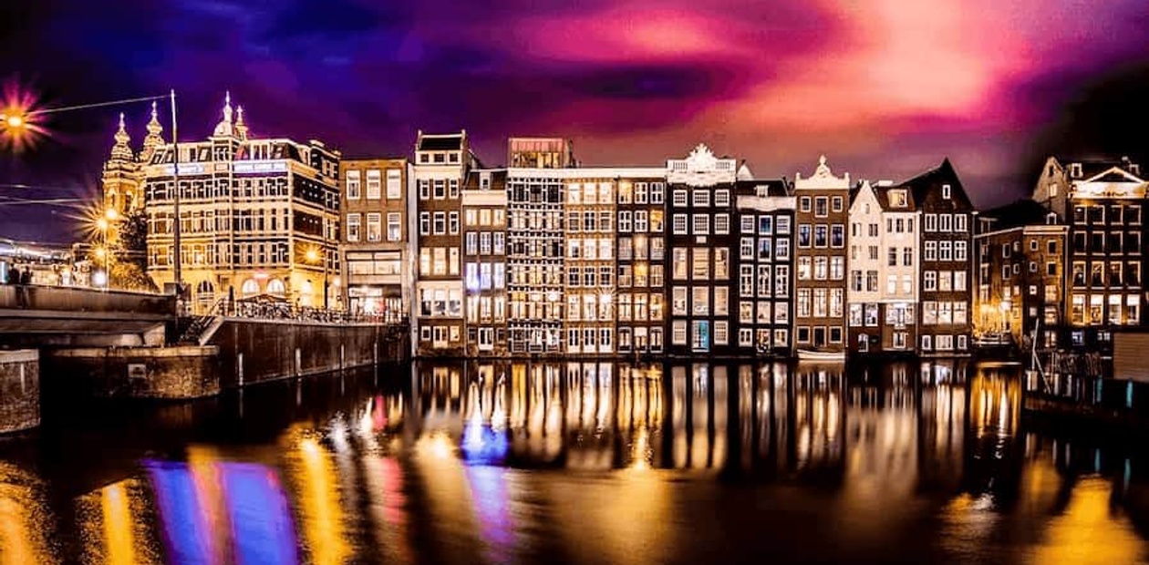 A colorful night view of Amsterdam with illuminated buildings reflected in the calm water of a canal.