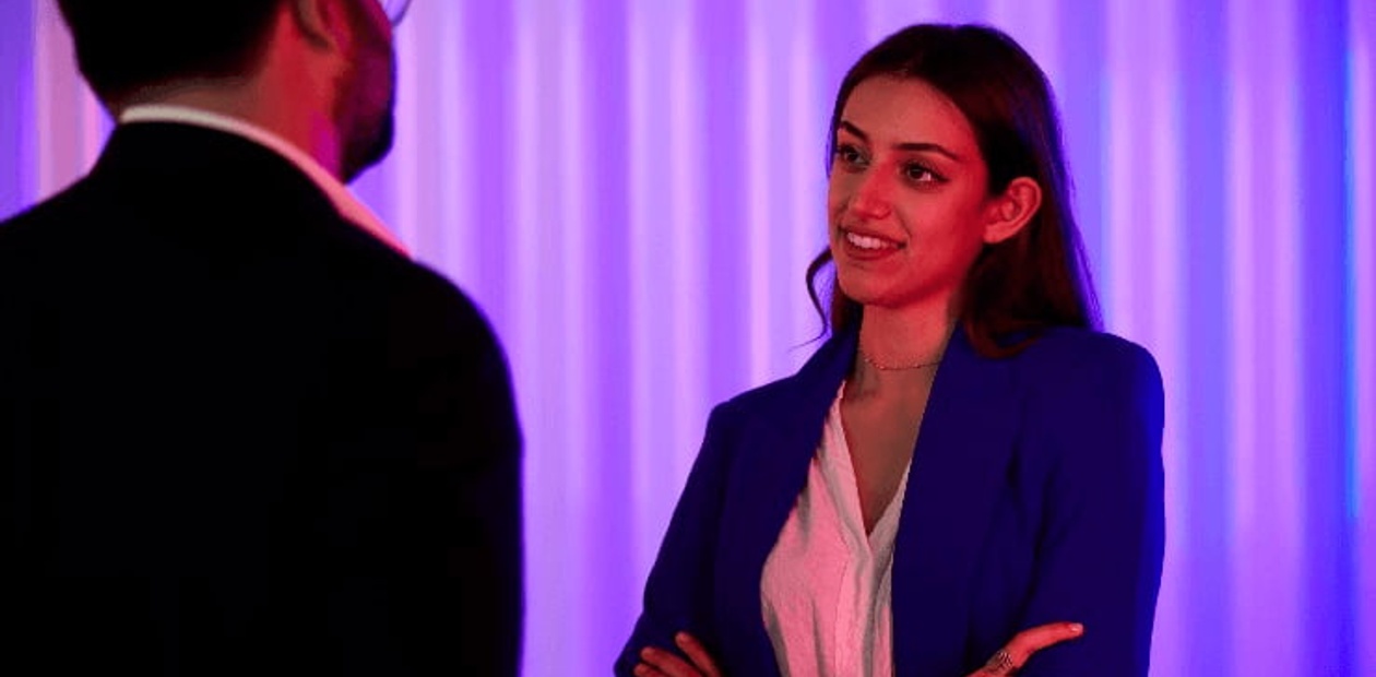 A woman in a blue blazer and white shirt talks to a man in a dark suit, with a purple background lighting.