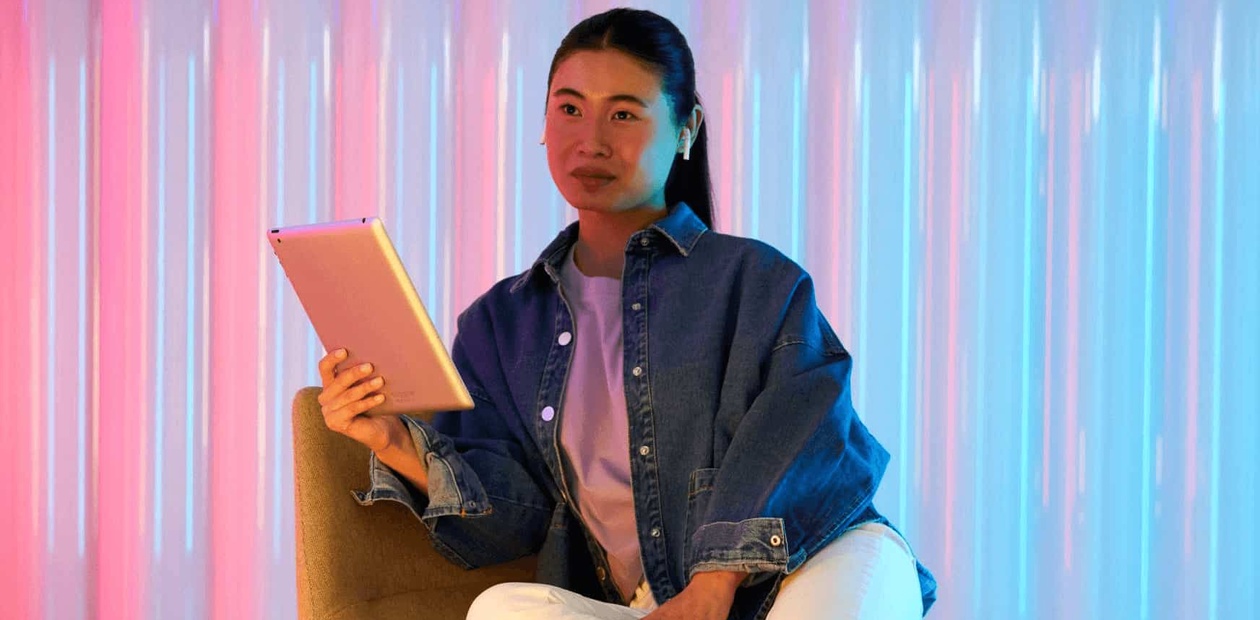 A woman sitting on a chair holding a tablet with a colorful neon light background.