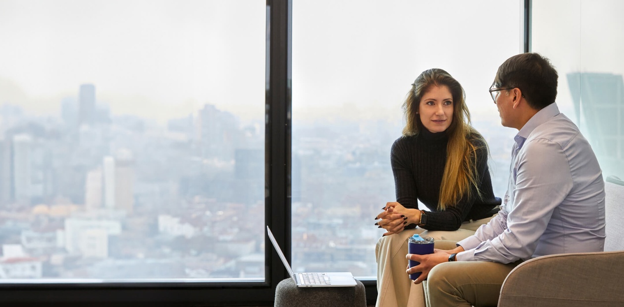 Two professionals engage in a conversation while sitting by a large window overlooking a cityscape.