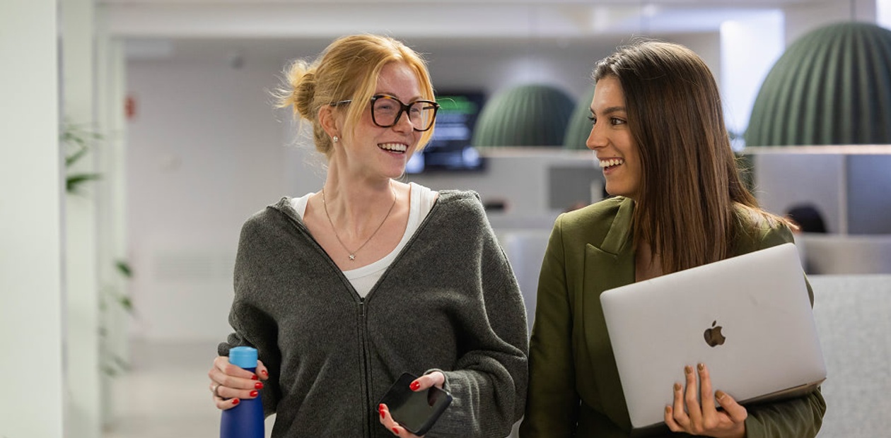 Two women are smiling and chatting while walking in a modern office environment.
