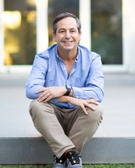 A smiling middle-aged man sitting casually on steps outdoors with a modern building in the background.