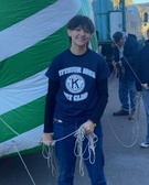 A young person holding ropes in front of a large green and white striped tent, wearing a black t-shirt with 'Preston Area Kiwanis Club' logo.