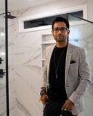 A man in a stylish gray blazer and black shirt standing in a modern bathroom with marble walls.