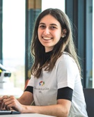 A young woman smiling at the camera while sitting at a desk with a laptop, wearing a white t-shirt with a 'Google Developer Clubs' logo.