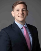 A professional headshot of a young man wearing a suit and pink tie, smiling at the camera.